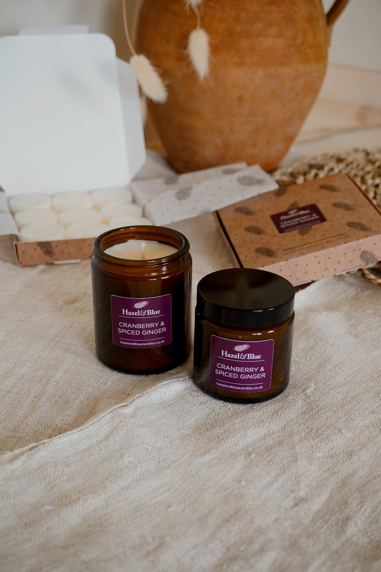 Two amber jar candles (180ml and 120ml) from our Autumn/Winter Collection with maroon/mauve labels, featuring the Cranberry & Spiced Ginger scent. Set in a cosy, autumnal scene, the warm glow evokes comfort, seasonal spice, and winter elegance.